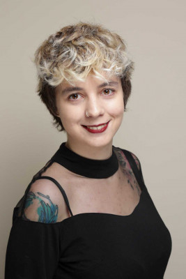 Ein Porträtfoto von Nadine vor einem einfarbigen, hellbeigen Hintergrund. Nadine lächelt leicht und ist von den Schultern bis zum Kopf zu sehen.

Kurze, lockige Haare mit dunklem Ansatz und hellblonden Strähnen oben. Helle Haut, braune Augen, dunkelroter Lippenstift. In der linken Nasenseite ein Nasenpiercing.

Getragen wird ein schwarzes Oberteil/Kleid mit halbtransparentem Mesh-Bereich an den Schultern und einem breiten, hoch sitzenden schwarzen Kragen. Auf der sichtbaren Schulter und am oberen Brustbereich sind farbige Tattoos zu erkennen (unter anderem blau/türkis auf der Schulter).